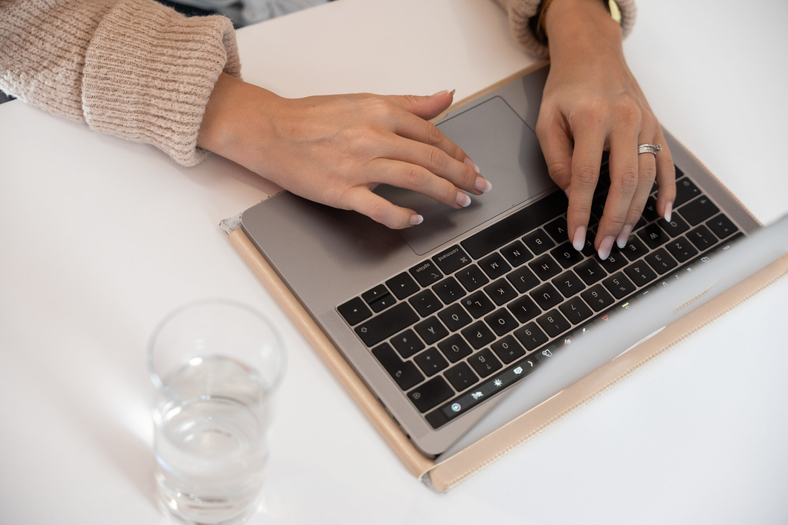 Hände einer Person, die einen Laptop auf einem weißen Schreibtisch verwendet, vielleicht um eine WordPress-Site zu erstellen oder Inhalte für SEO zu optimieren. Die Person trägt einen beigen Pullover und einen Ring, neben dem Laptop steht ein Glas Wasser.